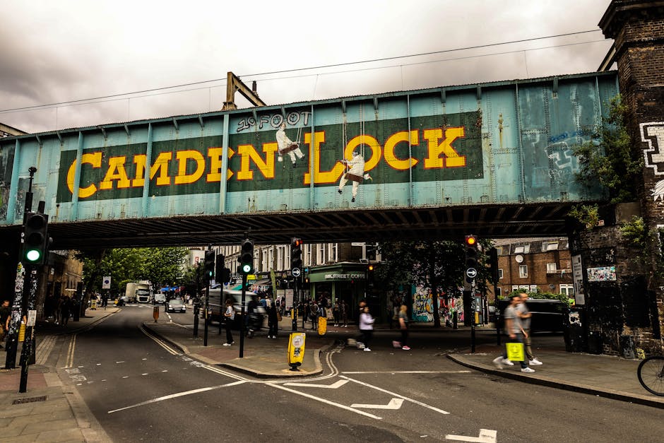 Exterior view of Camden Town Station with a red brick façade featuring large arched windows and a prominent blue sign displaying 'Camden Town Station'. The entrance is busy with numerous pedestrians,