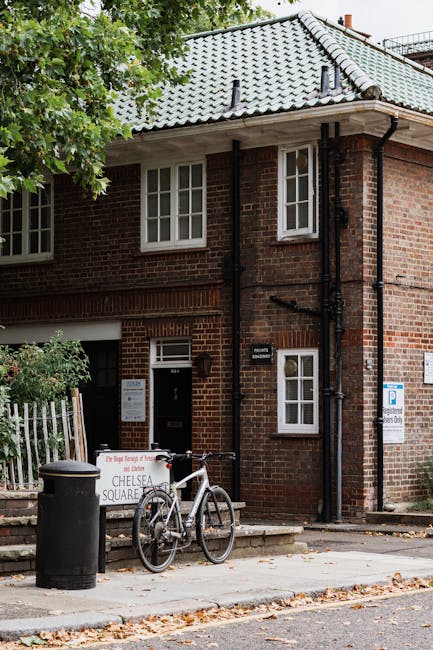 The exterior of a brick residential building in Chelsea SW3, featuring a tiled grey roof, white-framed windows, and a dark front door. A bicycle is parked against a small white fence near a black tras