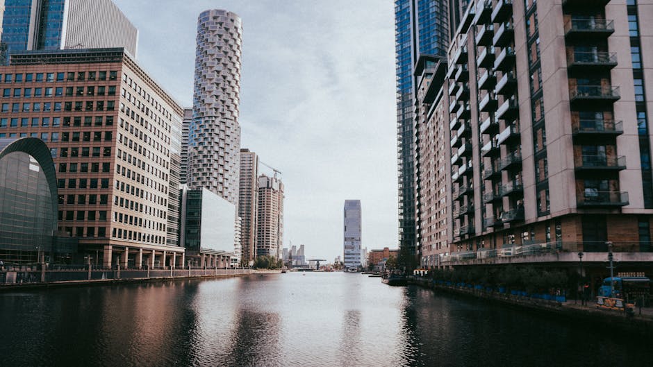 Photograph of a modern cityscape along a river, featuring tall commercial office buildings with glass and steel facades on both sides. The buildings include a range of architectural styles, some with 