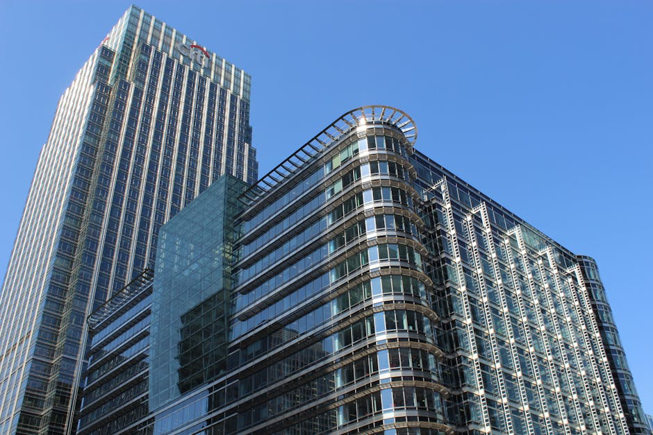 Photograph of a modern cityscape along a river, featuring tall commercial office buildings with glass and steel facades on both sides. The buildings include a range of architectural styles, some with 