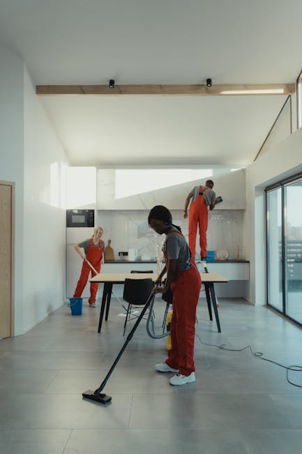 A team of professional cleaners wearing red aprons and casual clothing perform domestic cleaning tasks in a modern, minimalist kitchen with large glass doors allowing natural light. One cleaner in the