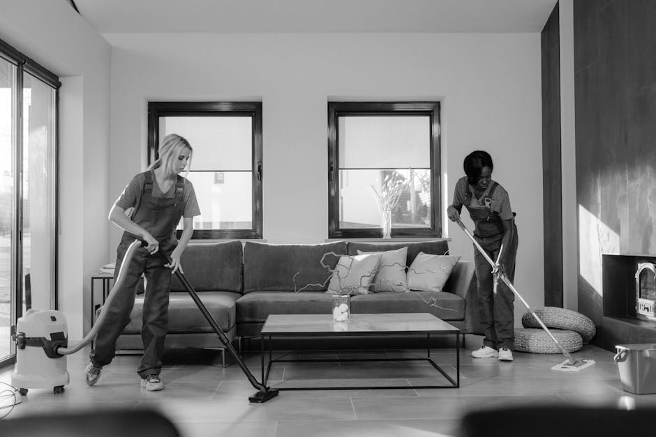 A team of professional cleaners wearing red aprons and casual clothing perform domestic cleaning tasks in a modern, minimalist kitchen with large glass doors allowing natural light. One cleaner in the